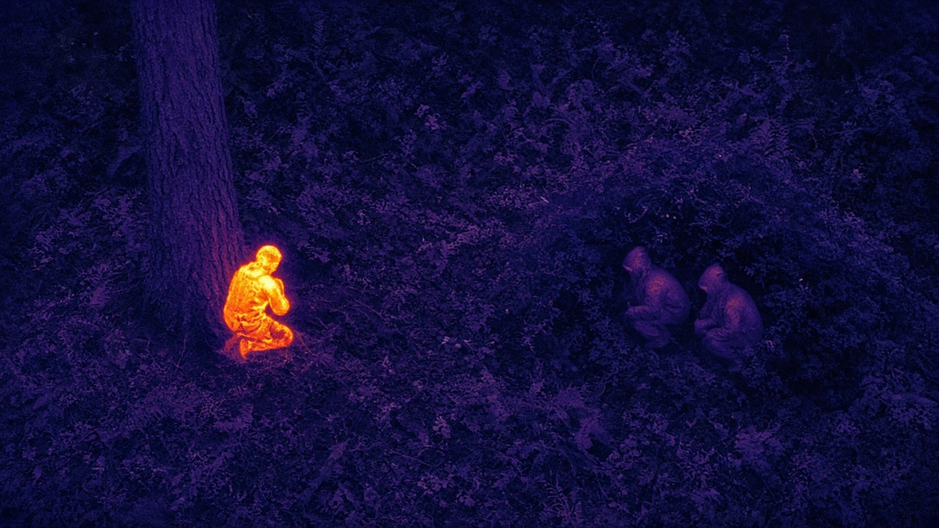 Drone thermal camera view of a forest at night – an uncovered soldier glows bright while two soldiers in green anti-thermal suits show only as faint cool shapes